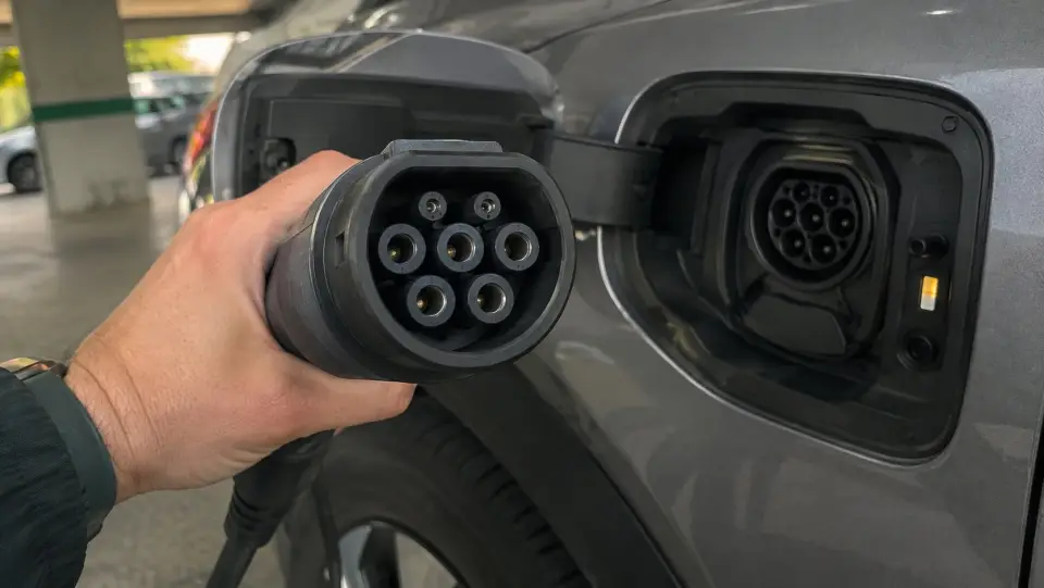 Mano sosteniendo un conector Tipo 2 de siete pines frente a la entrada de carga del auto